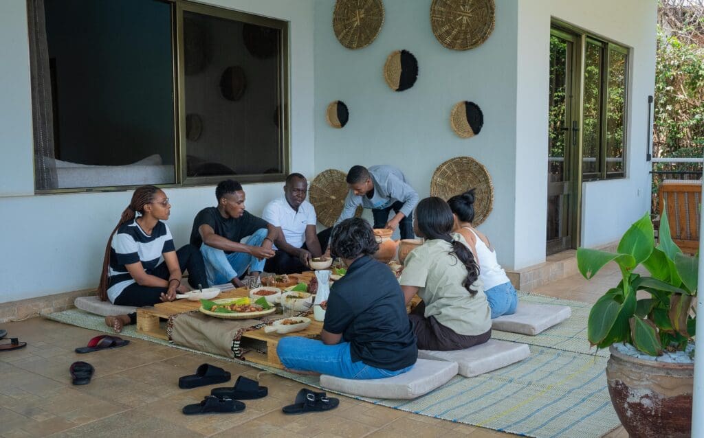 A group of people enjoying a traditional Rwandan meal together, seated around a low wooden table on a terrace at Judith Homestay.